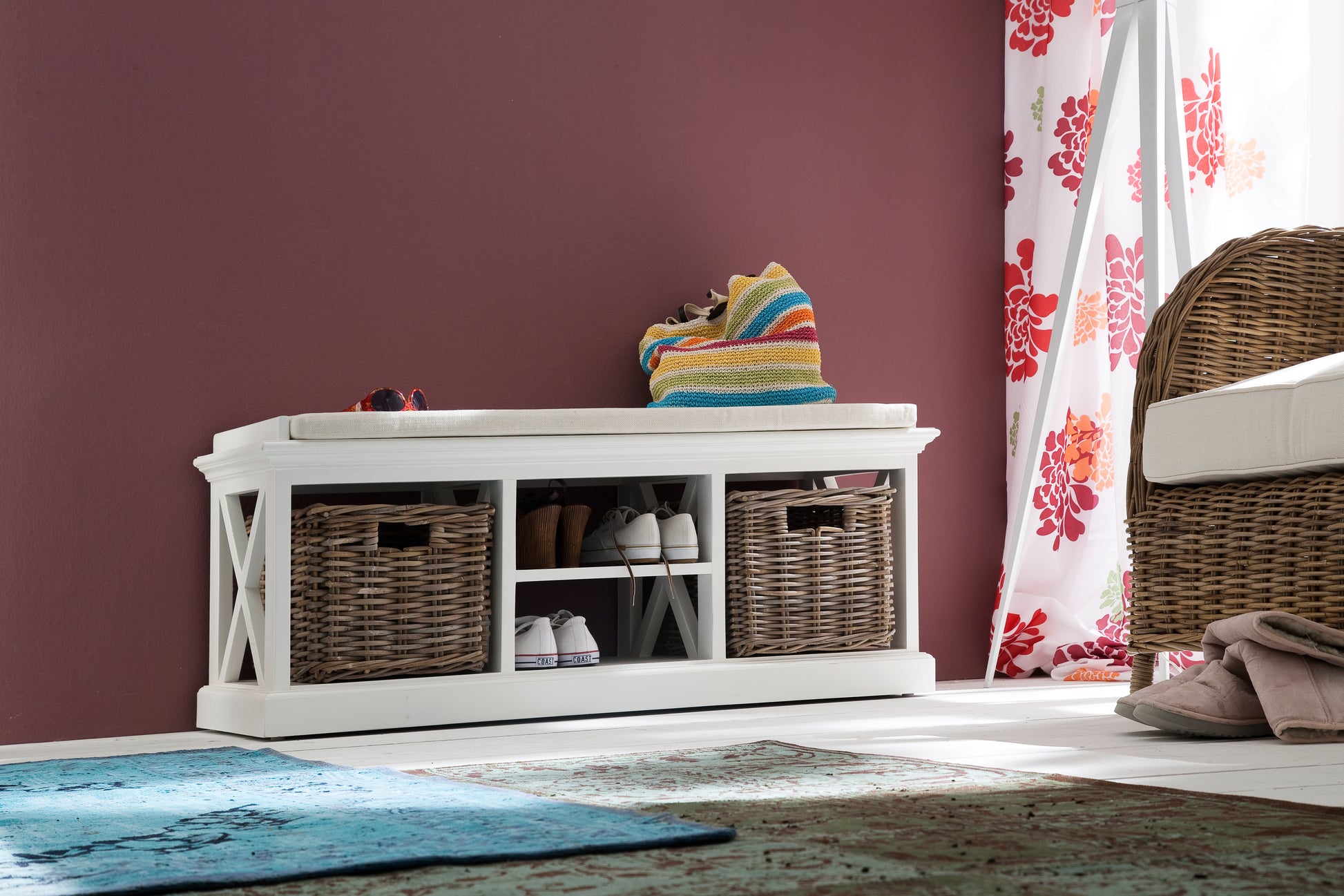 White timber bench with cushion and 2 basket set with shoes on the shelves next to a wicker armchair next to a bright window.