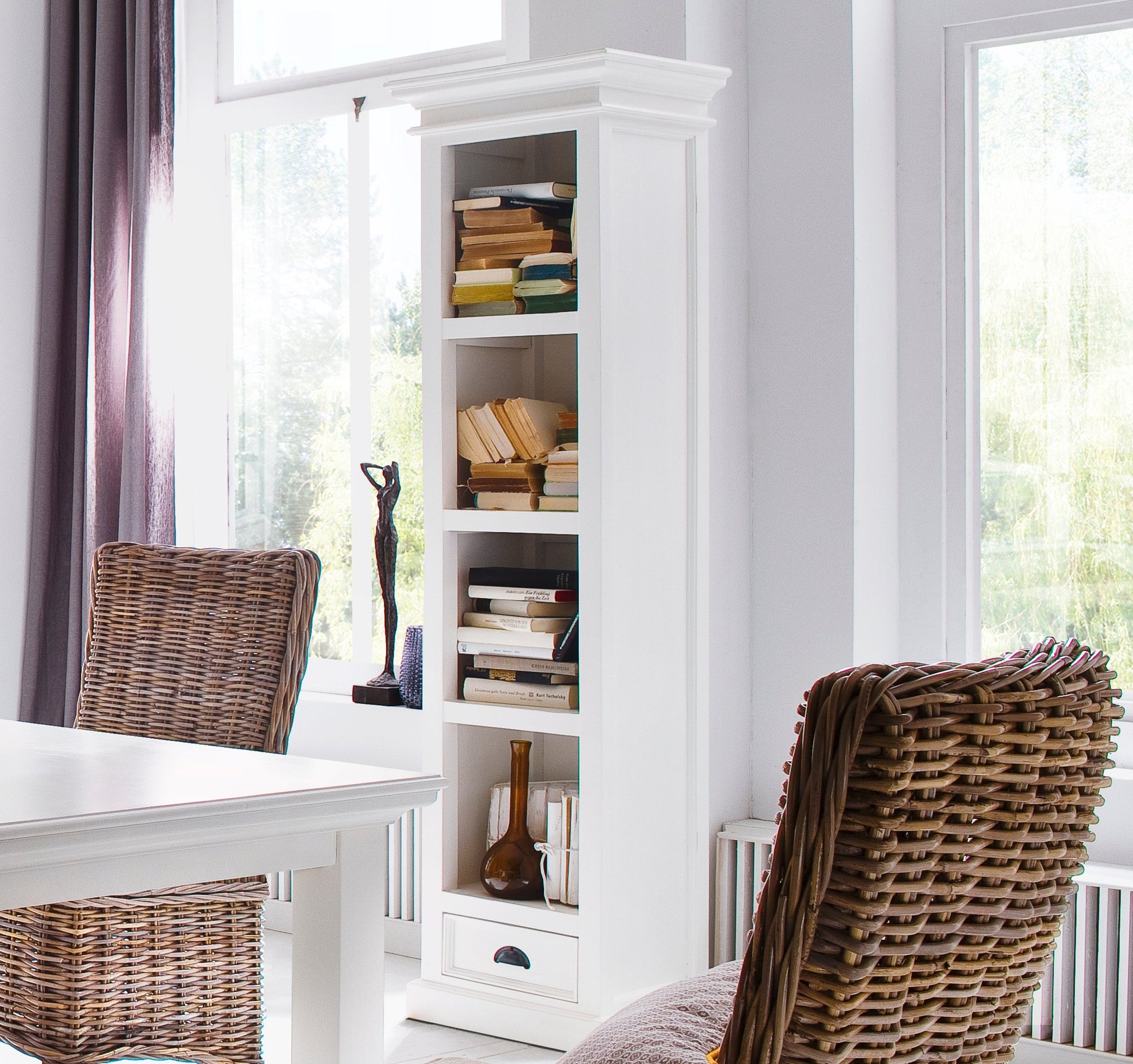 White wooden bookshelf with 4 shelves and 1 drawer with antique brass hardware next to a white dining table and wicker chairs.
