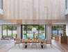 Wide interior shot of Andi Armchair Natural wooden dining chairs with a large dining table in an open-plan modern home, with floor-to-ceiling glass doors, pool view, pendant lighting, and minimalist Scandinavian architecture.