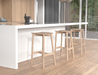 Close-up lifestyle view of Andi Backless Bar Stool - Natural - 66 cm Seat Height near a kitchen island, emphasising smooth curved seat edges, natural oak grain detail, and footrest support.