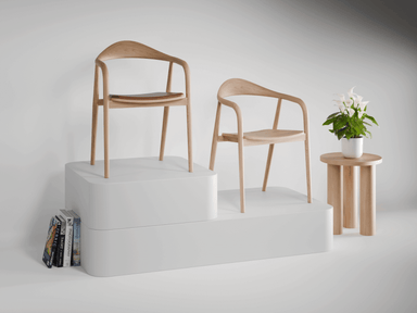 Two wooden armchairs in natural ash finish on a podium next to a small round wooden side table with a potted plant.