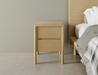 Simple natural wooden bedside table with two drawers beside a bed.