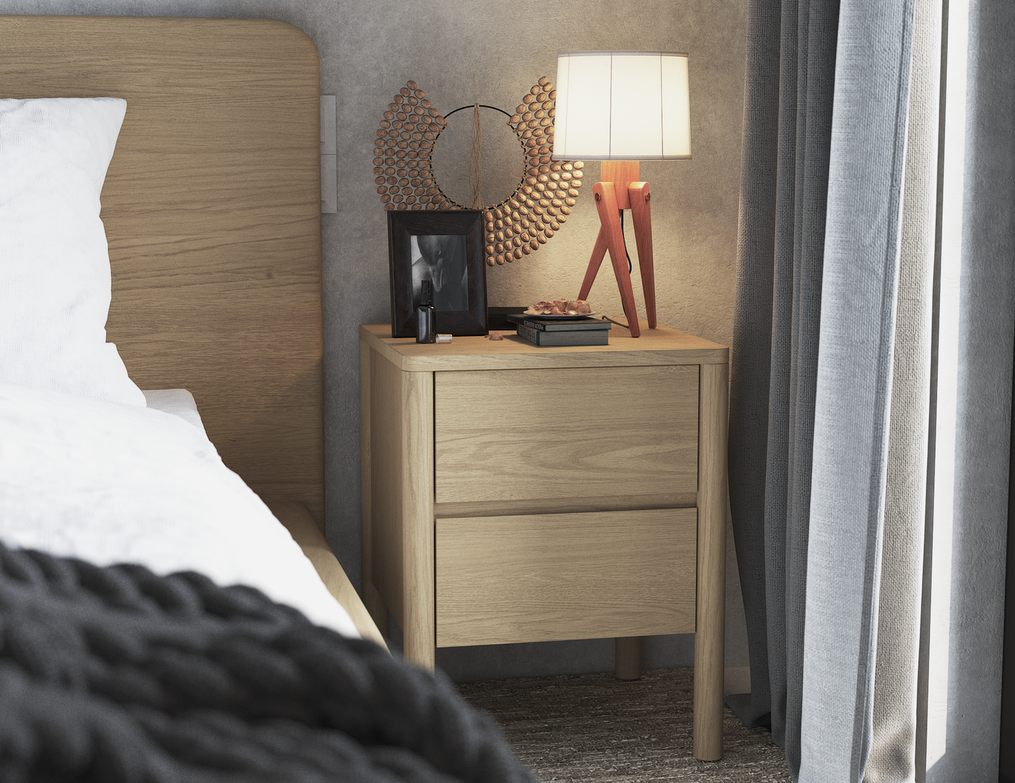 Natural wood bedside table with two drawers, next to a bed in a bedroom setting. The table is decorated with a modern lamp on a wooden tripod base, a round decorative mirror, a framed photo, and small personal items. 