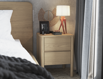 Natural wood bedside table with two drawers, next to a bed in a bedroom setting. The table is decorated with a modern lamp on a wooden tripod base, a round decorative mirror, a framed photo, and small personal items. 