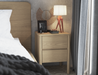 Natural wood bedside table with two drawers, next to a bed in a bedroom setting. The table is decorated with a modern lamp on a wooden tripod base, a round decorative mirror, a framed photo, and small personal items. 