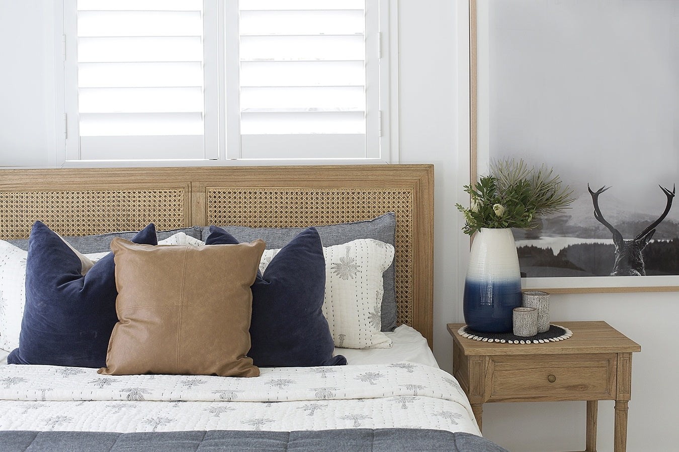 Hamilton Cane Bed Weathered Oak double bed styled bedroom scene with woven rattan cane headboard, white bedding, layered navy and tan cushions, timber bedside table, ceramic vase, and framed wall art in a bright modern interior.