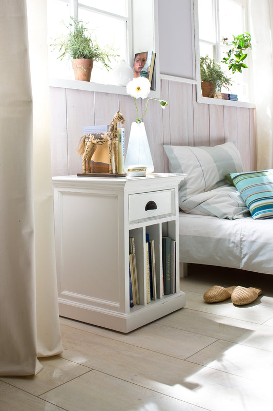 White timber bedside table with dividers, 1 drawer and antique brass hardware next to a bed in a bright bedroom setting.