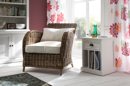 Casual setting of a white timber bedside table with dividers filled with books next to a wicker armchair and white timber buffet.