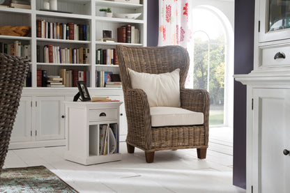 Home library setting with a white timber bedside table with dividers, a wicker armchair next to a large bright window and large book filled bookcase along the back wall.