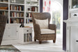 Home library setting with a white timber bedside table with dividers, a wicker armchair next to a large bright window and large book filled bookcase along the back wall.