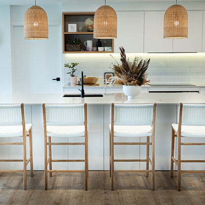 Wide lifestyle image of a bright kitchen with five Zen Counter Stools – White – Close Weave lined along a large island under pendant lights.