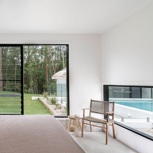 Interior bedroom setting with the Zen Armchair – Washed Grey – Open Weave beside large windows and a round timber side table.
