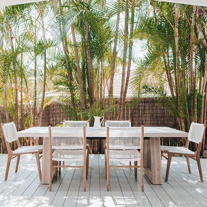 Outdoor patio scene with Zen Dining Chairs – White – Open Weave surrounding a large timber dining table under tropical palm shade.