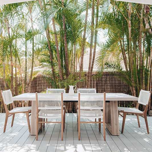 Outdoor patio scene with Zen Dining Chairs – White – Open Weave surrounding a large timber dining table under tropical palm shade.