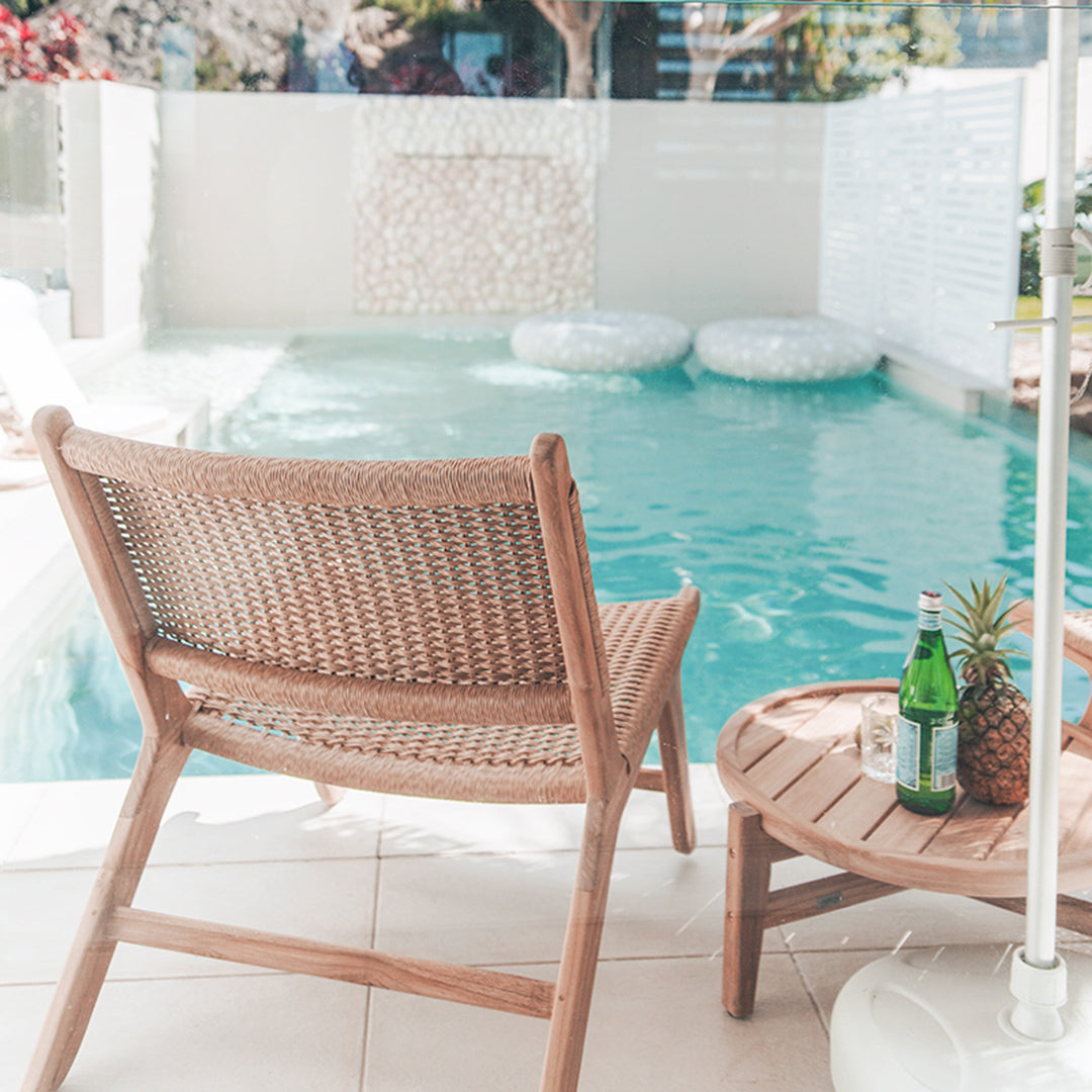 Rear view of Zen Lazy Chair – Sand – styled beside a circular teak table and tropical drink with pool water in background.