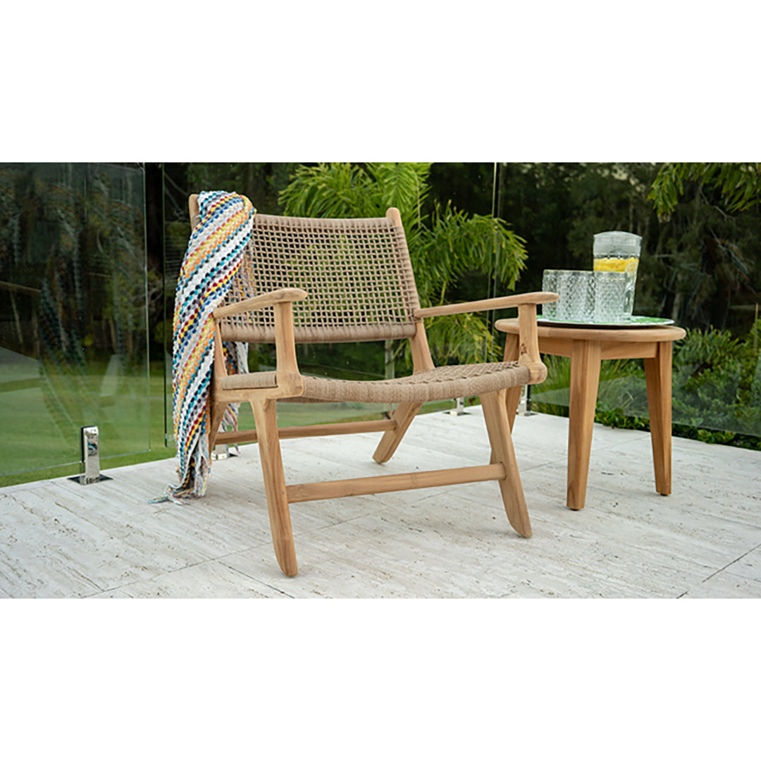 Outdoor scene featuring the Zen Lazy Chair - Sand - Open Weave on a light stone patio next to a round teak side table with water and lemon glasses. The chair has a teak frame with handwoven sand-coloured open-grid rope and a multicoloured throw draped over the backrest.