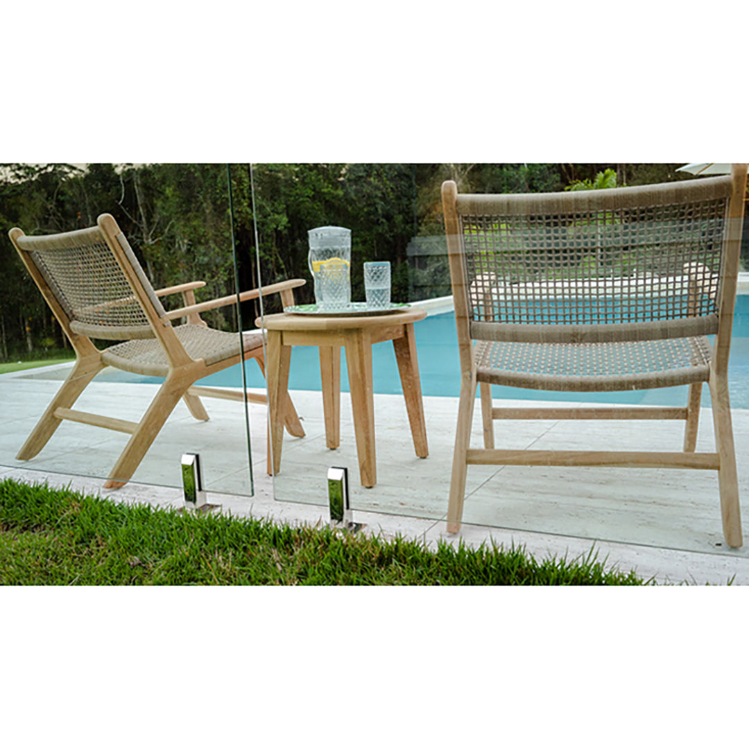 Poolside view of two Zen Lazy Chairs - Sand - Open Weave placed beside a round teak side table on a tiled patio. The chairs showcase sand-toned synthetic rope in an open-weave pattern on natural teak wood frames, with reflections visible in the glass fence.