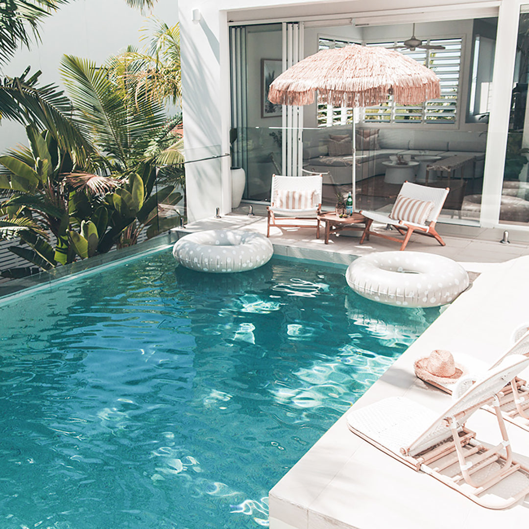 Poolside scene with two Zen Lazy Chairs - White - Open Weave arranged under a thatched umbrella, accompanied by a small teak table and striped cushions. The relaxed outdoor setting includes white pool floats and tropical greenery, highlighting the chair’s suitability for modern resort-inspired patios.