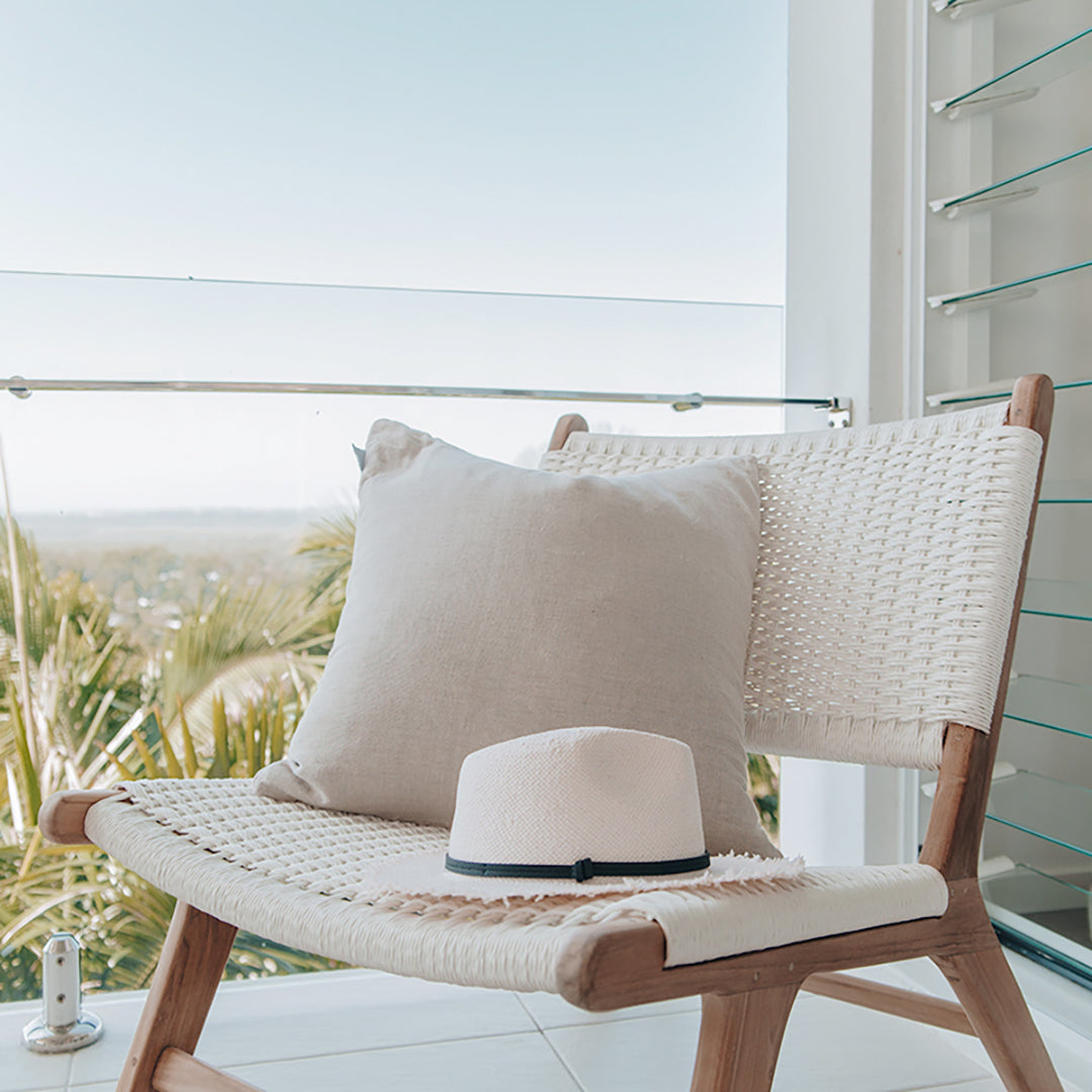 Close-up of Zen Lazy Chair on a balcony with a serene landscape view, featuring a soft beige throw pillow and a white summer hat resting on the seat. The white open weave and teak wood structure evoke a tranquil, resort-style outdoor escape.