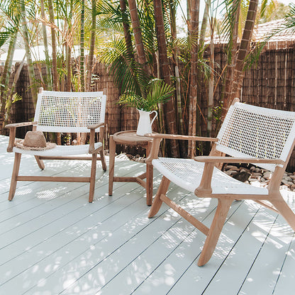 Two Zen Lazy Chairs - White - Open Weave styled outdoors on a sunlit wooden deck surrounded by palm trees and a bamboo privacy fence. Each chair features a handwoven white open-weave rope seat and back on natural teak wood frames, arranged with a round teak side table holding a white ceramic jug and a palm frond, evoking a relaxed tropical ambiance.