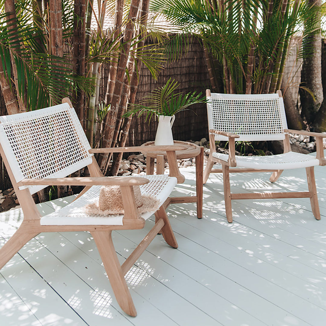 Two Zen Lazy Chairs - White - Open Weave styled outdoors on a sunlit wooden deck surrounded by palm trees and a bamboo privacy fence. Each chair features a handwoven white open-weave rope seat and back on natural teak wood frames, arranged with a round teak side table holding a white ceramic jug and a palm frond, evoking a relaxed tropical ambiance.