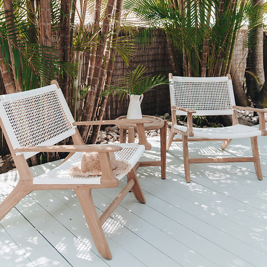 Two Zen Lazy Chairs - White - Open Weave styled outdoors on a sunlit wooden deck surrounded by palm trees and a bamboo privacy fence. Each chair features a handwoven white open-weave rope seat and back on natural teak wood frames, arranged with a round teak side table holding a white ceramic jug and a palm frond, evoking a relaxed tropical ambiance.