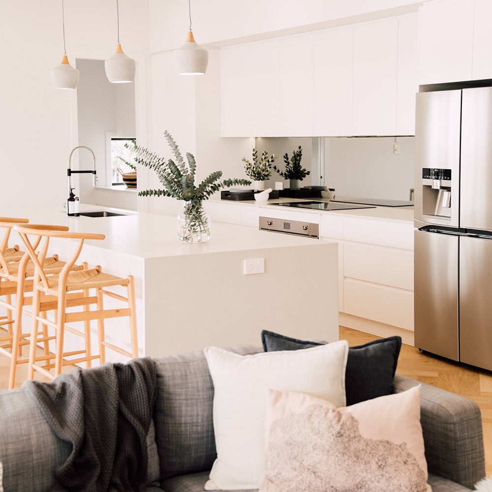 Wishbone Counter Stool in Natural Oak in an open plan kitchen and living room with white island bench, grey sofa and coastal home styling.